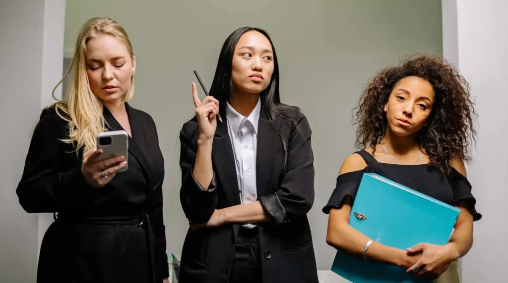 Current image: Three diverse women in black office wear standing against a gray wall, each holding office supplies